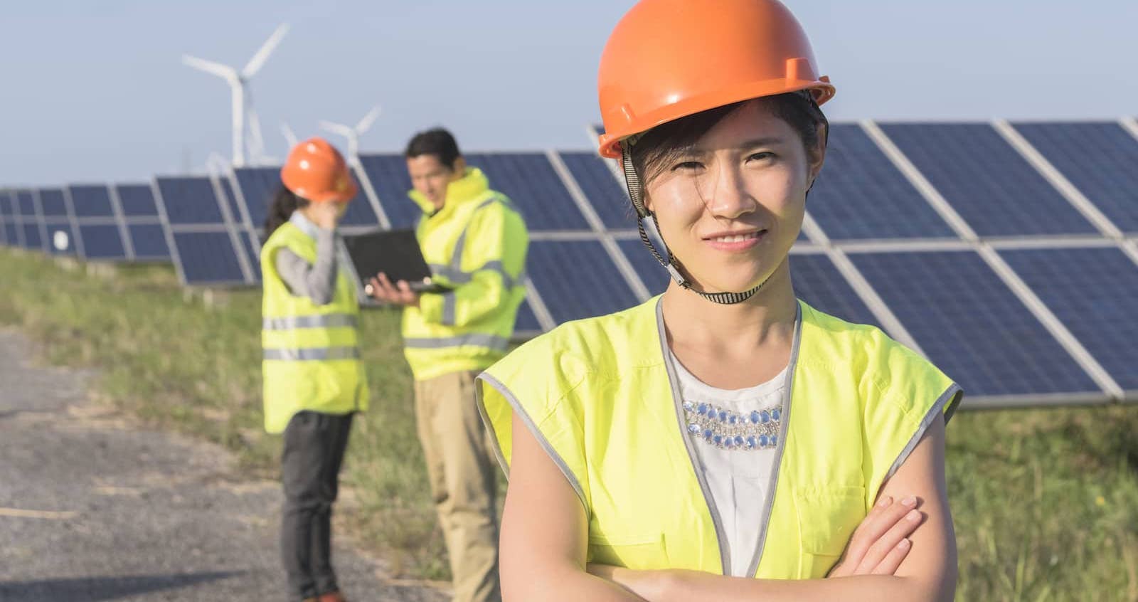 A woman in a hard hat smiling at the camera with two others talking in the background